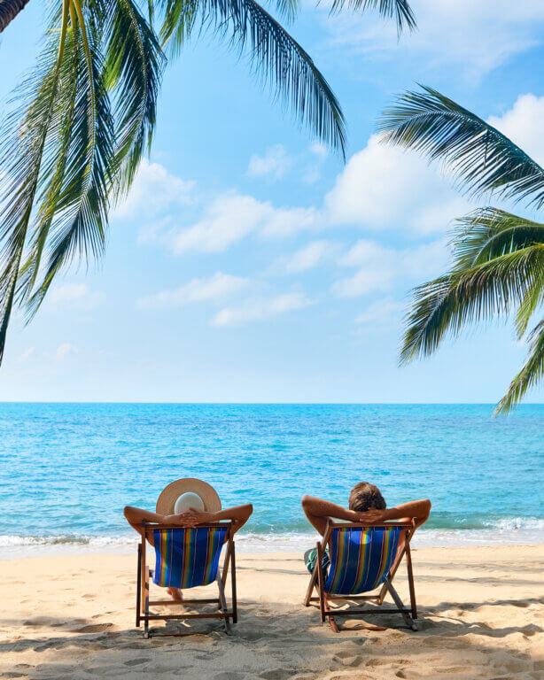 Two striped beach chairs facing the ocean under palm trees on a sandy beach.