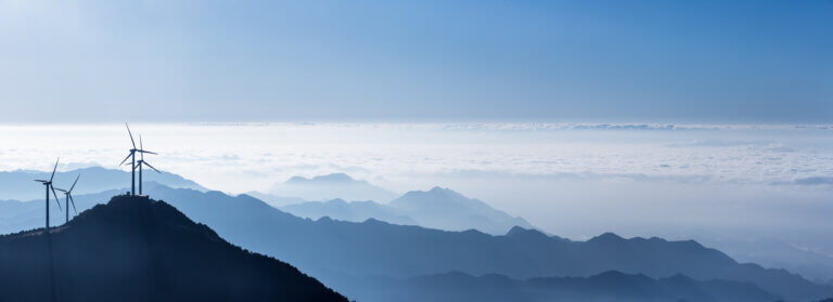 Wind turbines positioned on a mountain ridge above a layer of clouds with distant mountains in the background.