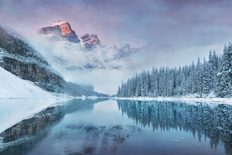 Snow-covered mountains reflecting in a calm lake surrounded by evergreen trees.