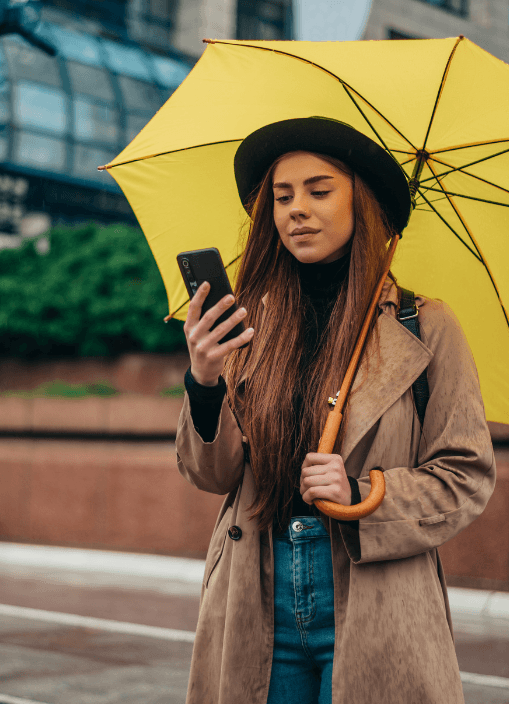 Woman with long brown hair wearing a beige trench coat and black hat holds a bright yellow umbrella while scrolling on a smartphone in an urban setting.