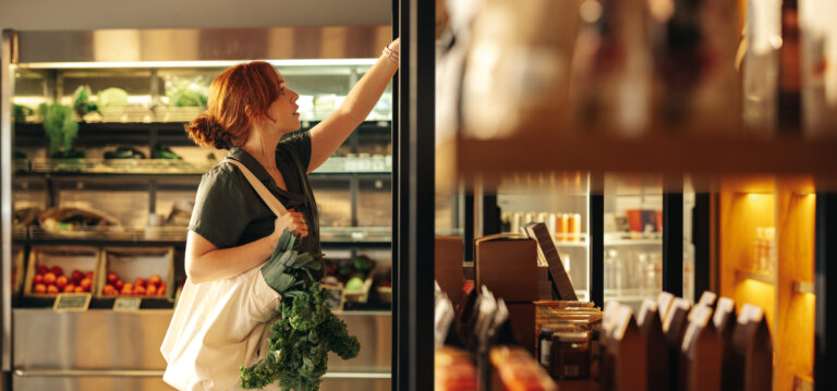 Woman with red hair reaching for items on a shelf in a grocery store while holding a bag of kale.