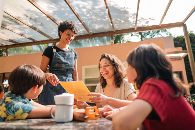 A waitress presenting a menu to a family seated at a table outdoors in a restaurant.