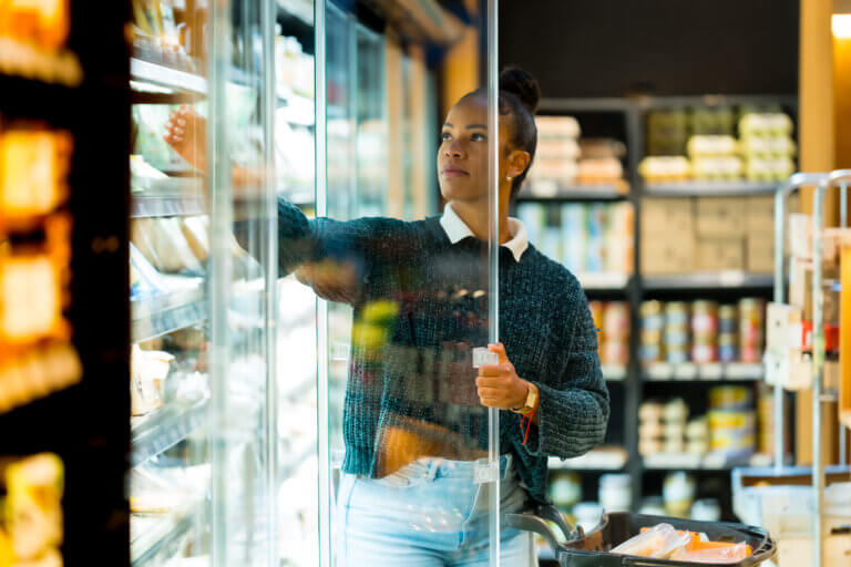 Woman in a grocery store reaching for products in a freezer section while holding a shopping cart.