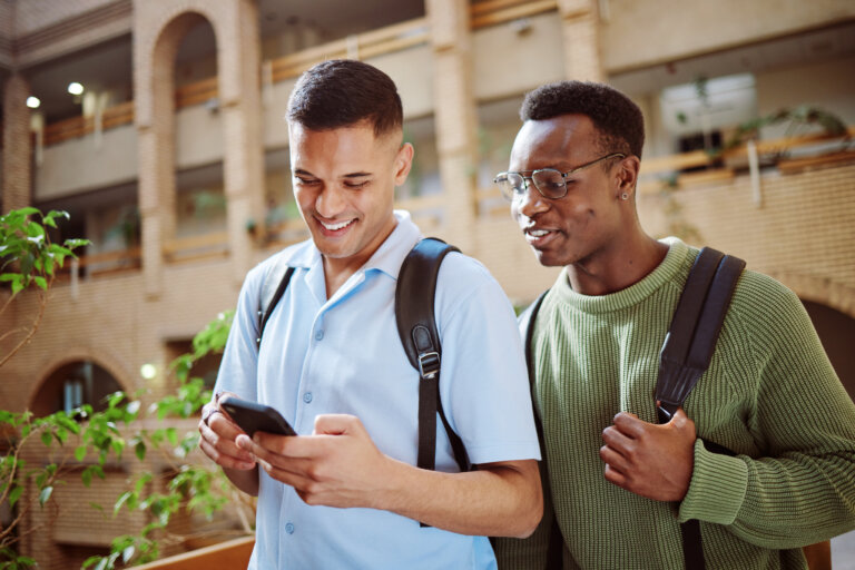 Two young men standing together, one looking at a smartphone while the other observes with interest.