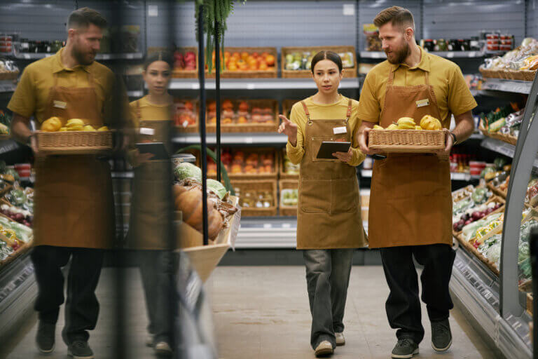Two grocery store employees, one carrying a basket of produce, discussing in an aisle filled with fruits and vegetables.